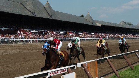 The grandstand at Saratoga Race Course in the background, with four horses in a line running towards the finish line beneath a clear blue sky